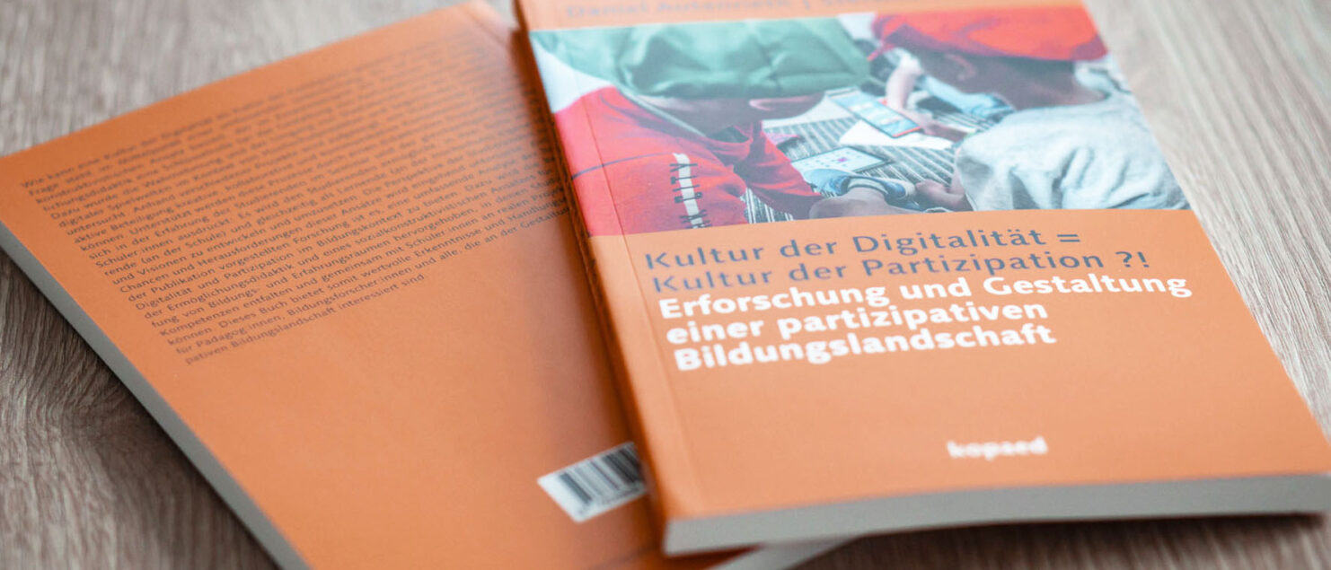 Vorder- und Rückseite von zwei Büchern "Kultur der Digitalität = Kultur der Partizipation ?! Erforschung und Gestaltung einer partizipativen Bildungslandschaft" von Autenrieth & Nickel, auf einem Holztisch.